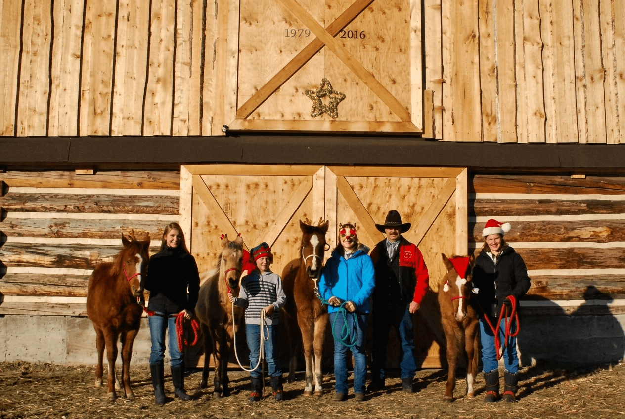family-and-horses-christmas-photo-at-barn A family posing with festive horses outside a wooden barn.
