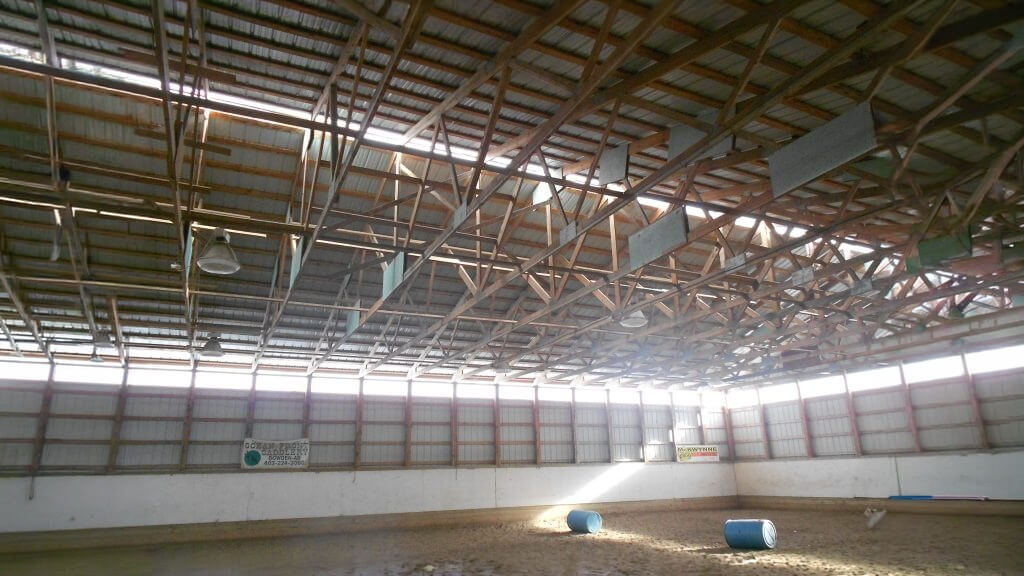 indoor-riding-arena Empty indoor riding arena with high wooden truss ceiling and blue barrels on the sand floor.