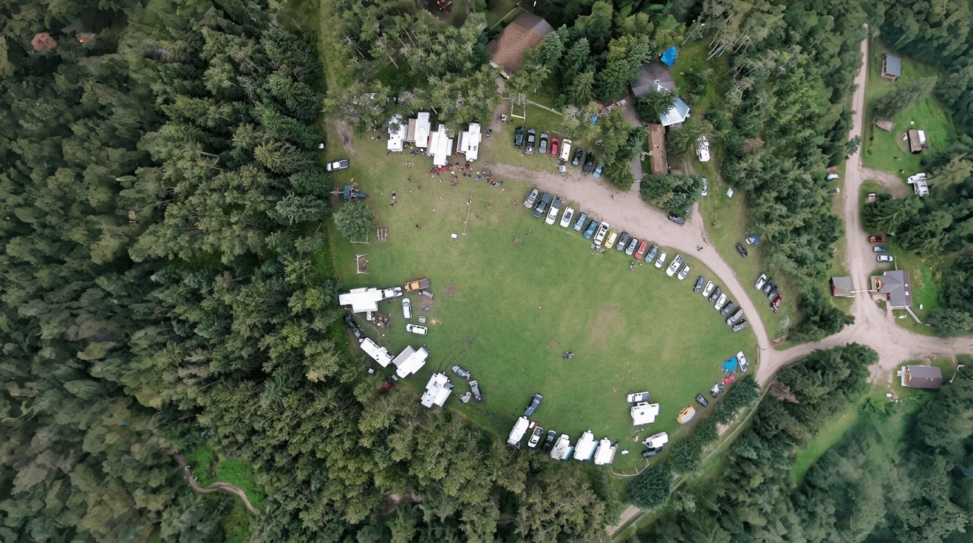 overview_room to roam Aerial view of a campground with RVs and vehicles parked in a grassy clearing surrounded by forest.