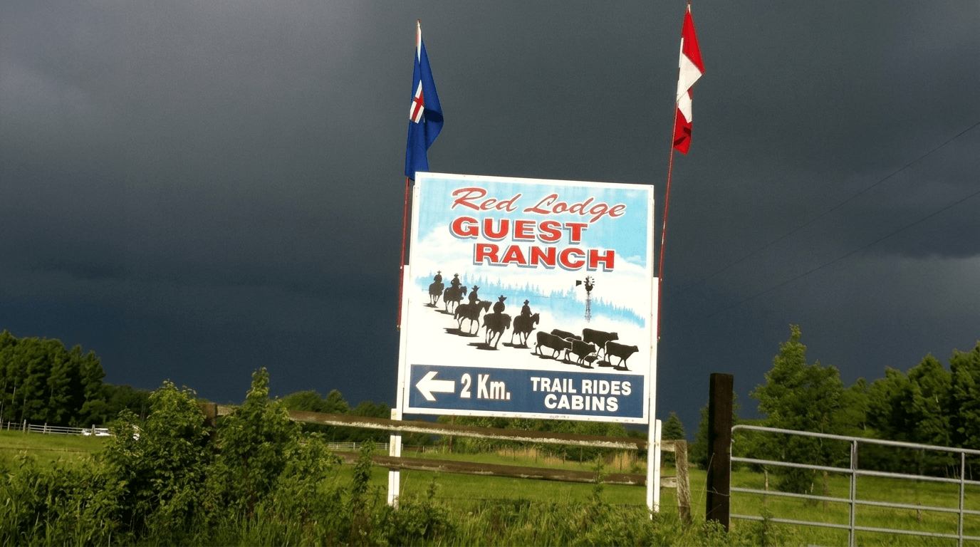 red-lodge-guest-ranch-sign-with-riders Outdoor sign for Red Lodge Guest Ranch showing horseback riders and cattle under a dark sky.