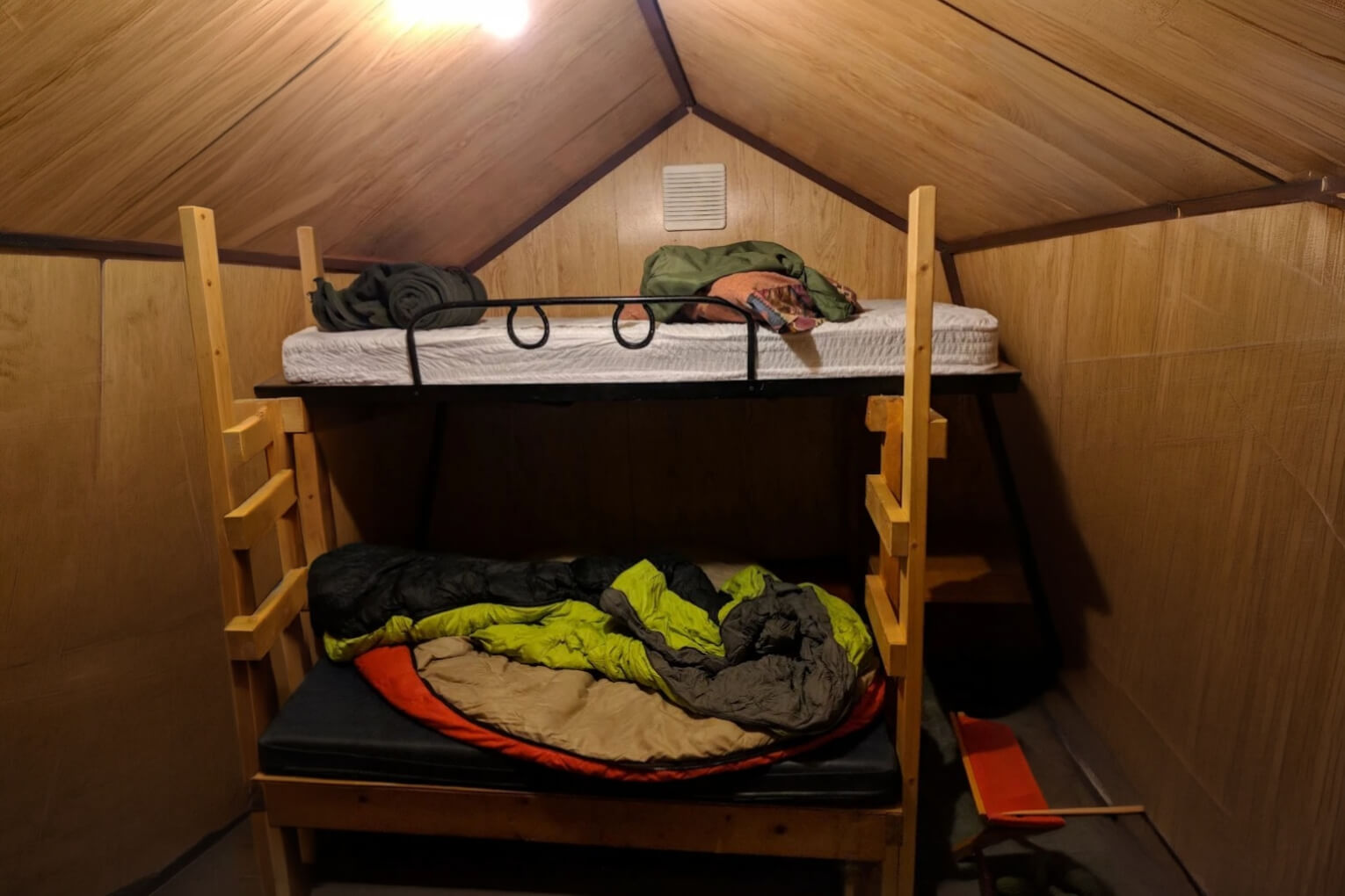 small cabin A cozy wooden bunk-bed cabin with luggage and a sleeping bag on the upper bunk, and a person tucked under a green blanket below; warm, quiet mood.