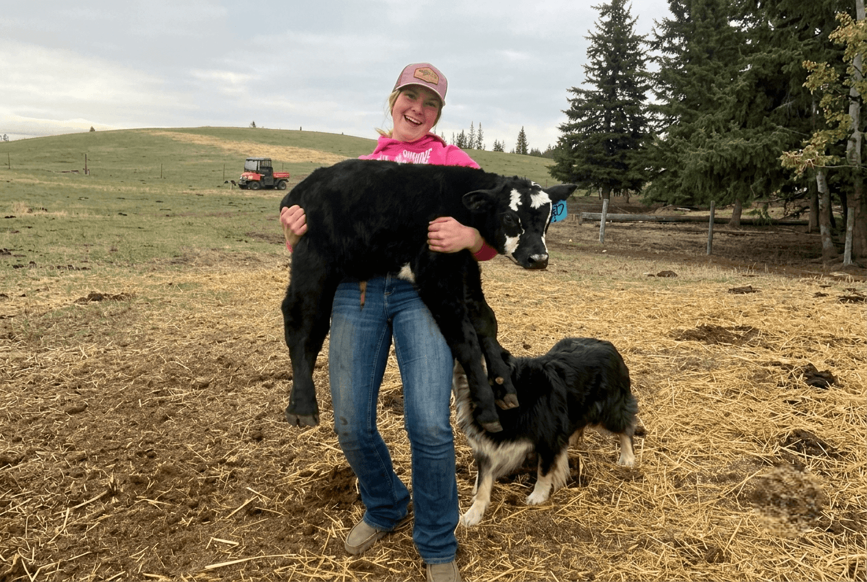 woman-holding-black-calf-with-dog A woman carrying a small black calf next to a farm dog.