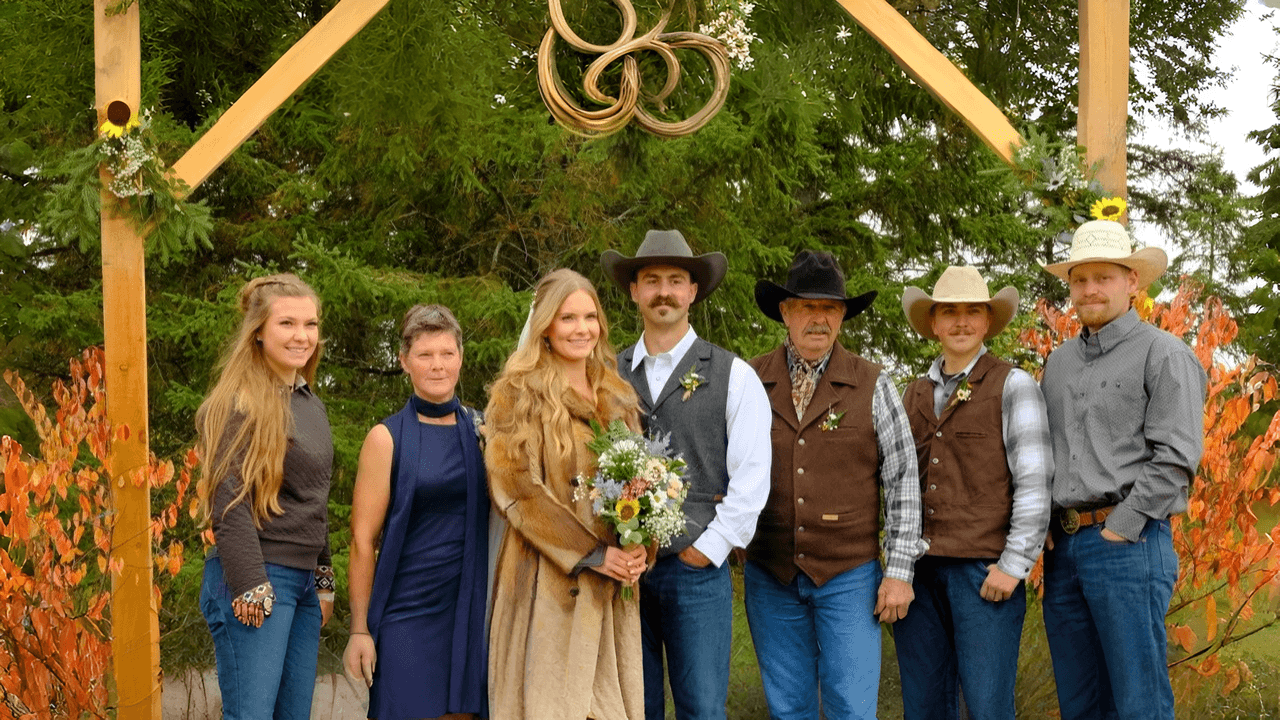 Wedding party poses under a wooden arch with autumn foliage and evergreen trees.